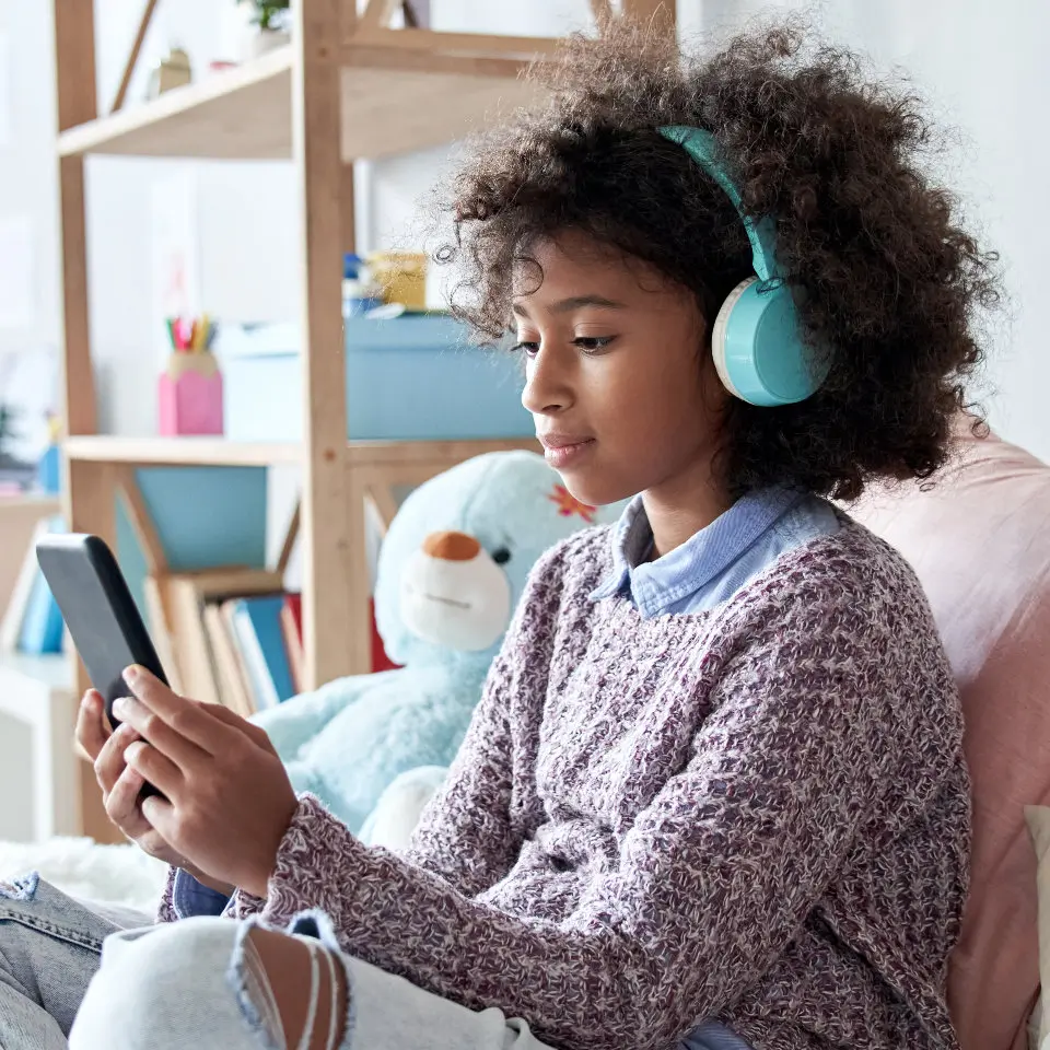 A young girl sits on a bed, wearing headphones and looking at her smartphone. A stuffed teddy bear is beside her.