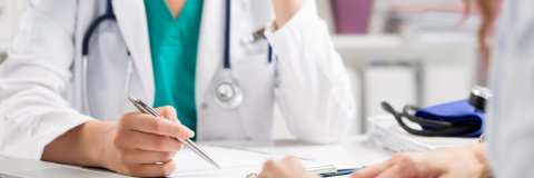 Two medical professionals in white coats discussing documents at a desk. One holds a pen, the other has a stethoscope around their neck.