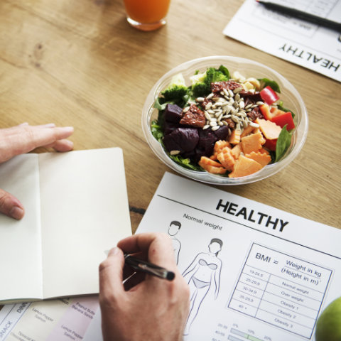 Person writing in a notebook next to a salad bowl, BMI chart, and a paper labeled "Healthy" on a wooden table.