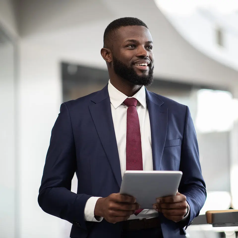 A man in a suit holds a tablet while smiling in a modern office setting.
