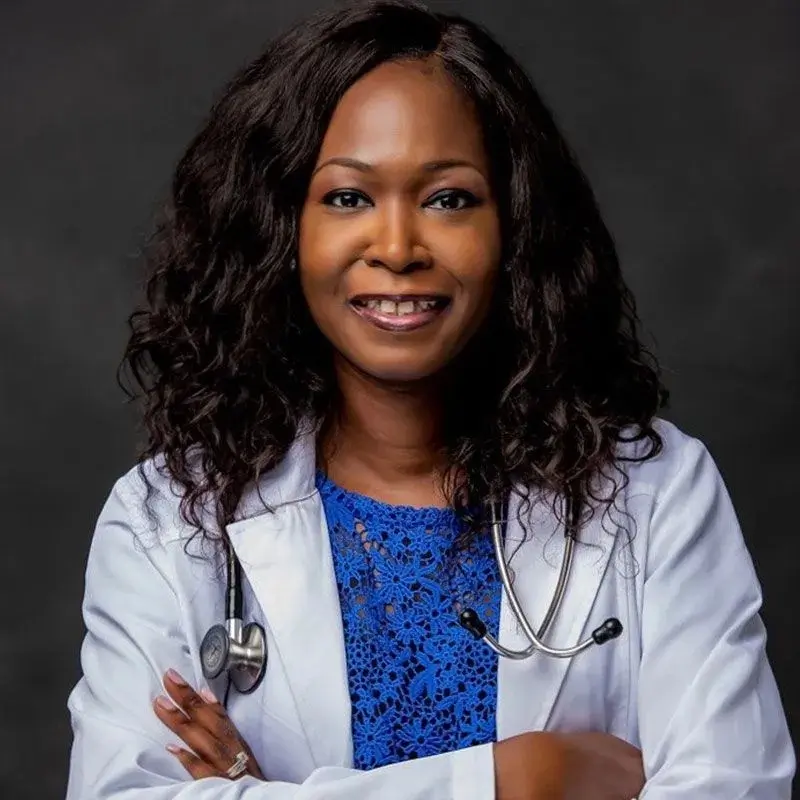 Person smiling, wearing a white lab coat and stethoscope, with arms crossed against a dark background.