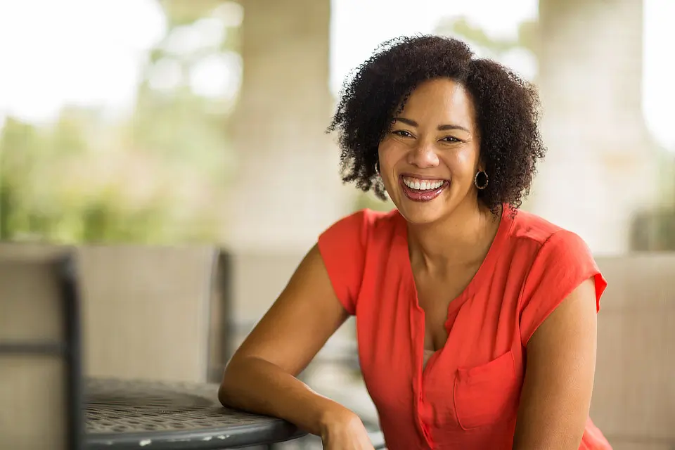 A person wearing a coral blouse sits at a table outdoors, smiling at the camera.