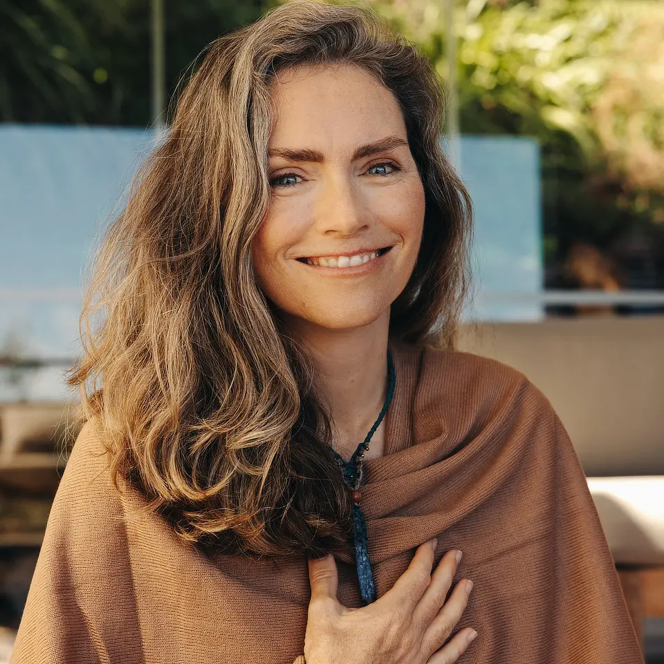 Woman with long hair and a shawl smiles at the camera, with greenery in the background.