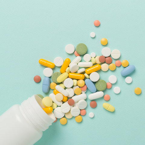 A variety of multicolored pills and tablets spill from a white bottle onto a light blue background.