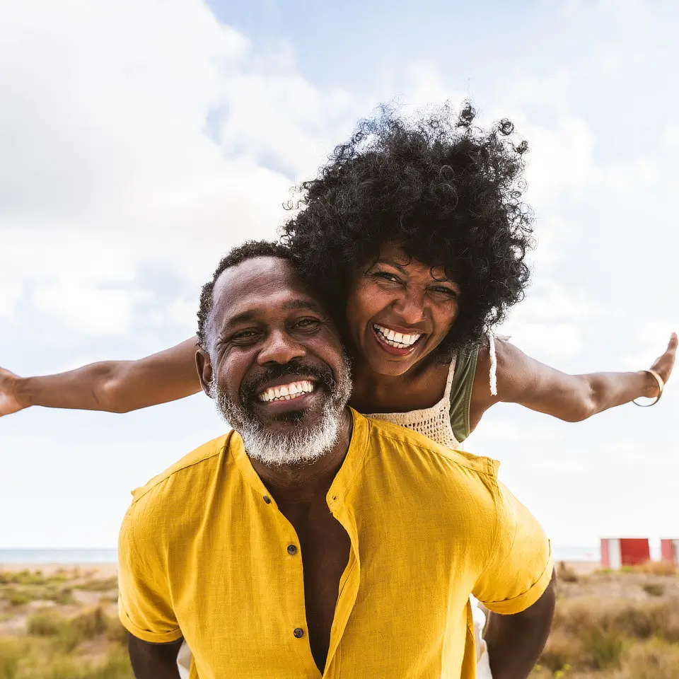 Man in yellow shirt giving a piggyback ride to a joyful woman with curly hair outdoors, both smiling.