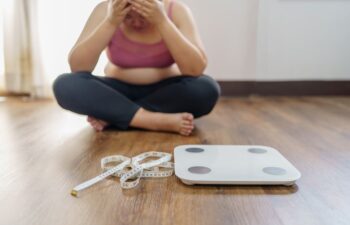 Upset obese woman wearing sports clothes sitting on the floor next to the scales and measuring meter hiding her face in her hands