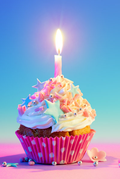 A cupcake with white and pink icing, decorated with star-shaped sprinkles and a lit candle, sits in a pink polka dot wrapper against a blue and pink gradient background.