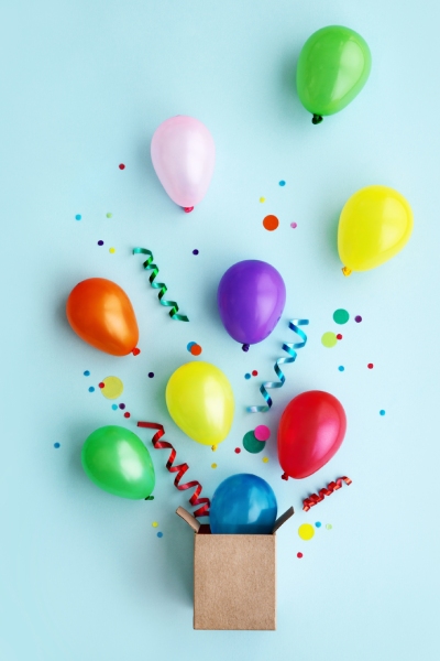 A small cardboard box with colorful balloons, streamers, and confetti coming out, set against a light blue background.