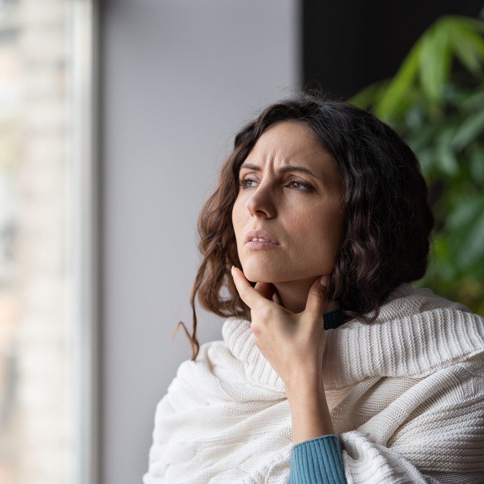 Upset sick woman touching painful swollen neck glands, suffering from throat infection, unhealthy female standing near window at home feeling pain with swallowing, selective focus